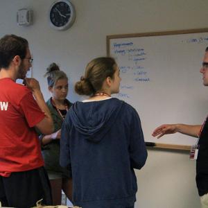 Three students and an instructor have a discussion in front of a whiteboard