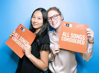 Emily Abshire and another NPR intern post with orange posters that read "All Songs Considered"
