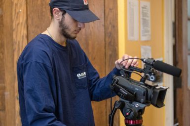 Media School sophomore Anthony Rodman stands behind a video camera and looks into it.