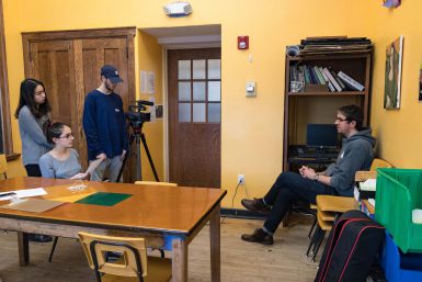 Media School freshman Jiatong Zou, Harmony Middle School eighth grader Julie Mendez and Media School sophomore Anthony Rodman gather behind a camera, which points at Hugh Farrell, a staff member at Community Kitchen of Monroe County.