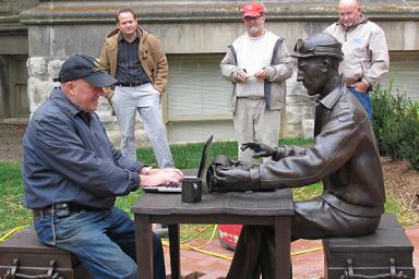 Sculptor Tuck Langland takes a seat on a munitions crate opposite Ernie Pyle. Langland left space on the table for visitors to work on their laptops.