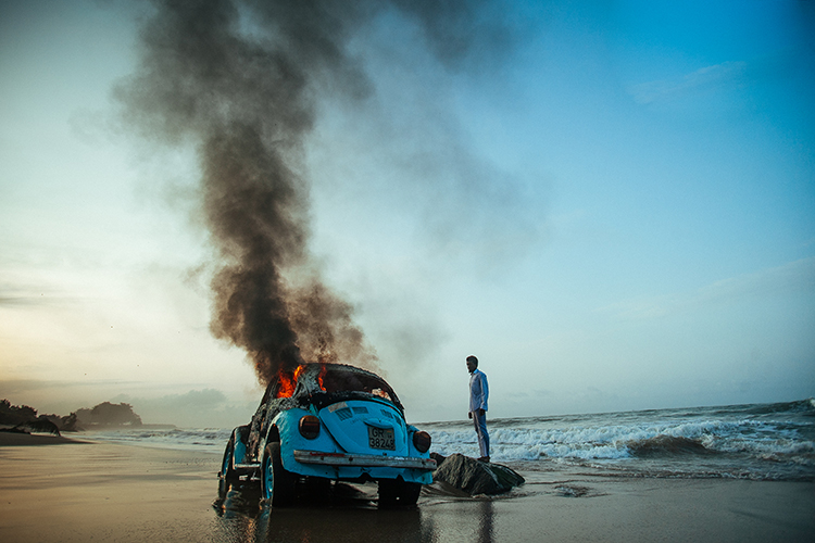 A blue Volkswagon Beetle on fire on a beach. A man watches.