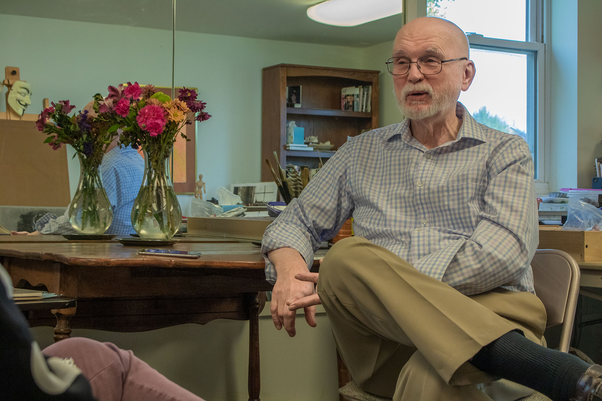 Professor emeritus Claude Cookman sits in a chair next to a table with a vase of flowers, in front of a mirror.