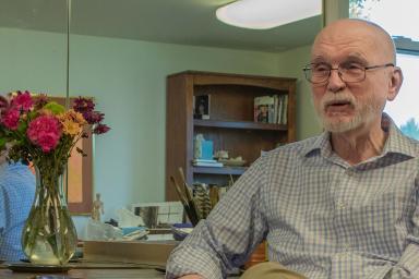 Professor emeritus Claude Cookman sits in a chair next to a table with a vase of flowers, in front of a mirror.