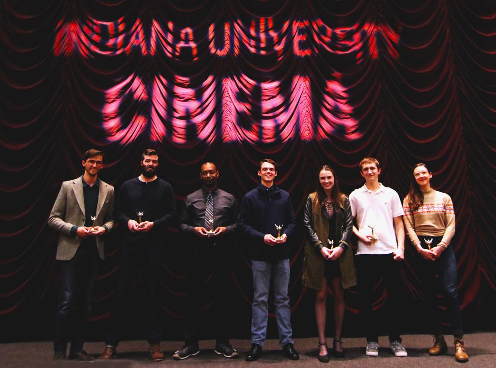 Seven students pose in front of a red theater curtain holding trophies