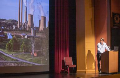 Balog watches from a lectern as an image of a city with smokestacks appears on the screen.