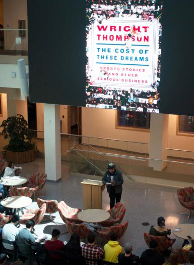 Wright Thompson speaks at a podium in front of a large digital screen. The screen displays an image of his book. The text says, "Wright Thompson: The Cost of These Dreams: Sports Stories and Other Serious Business." The text is on top of an image of boxers in a ring.