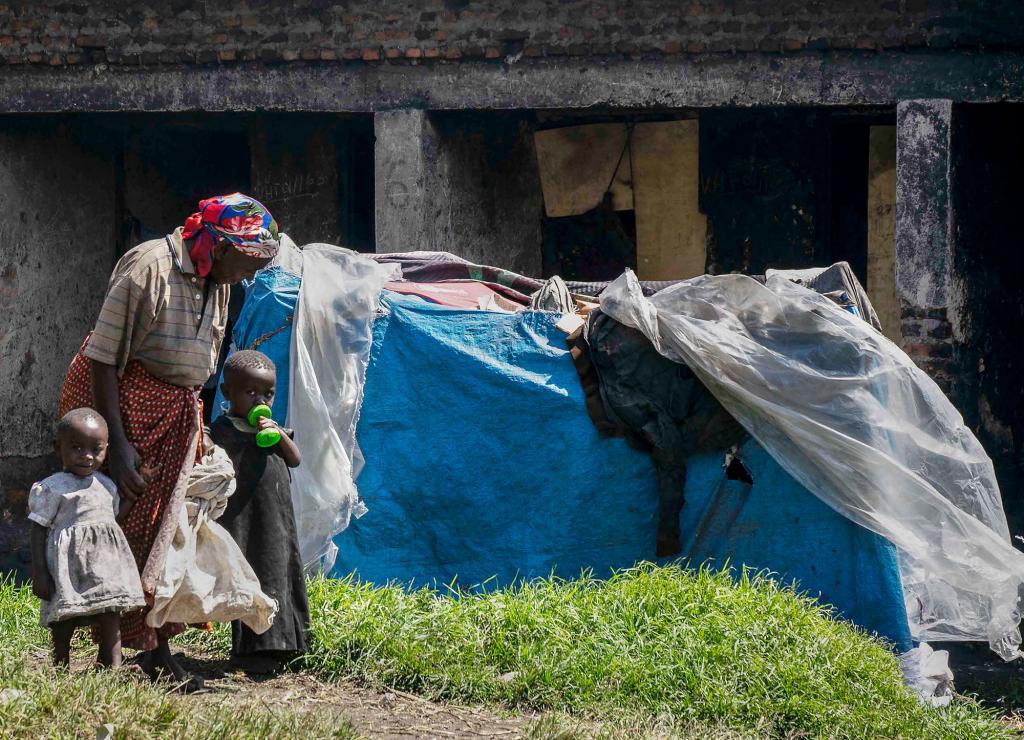 A mother and two children stand next to a tent made of plastic sheeting.