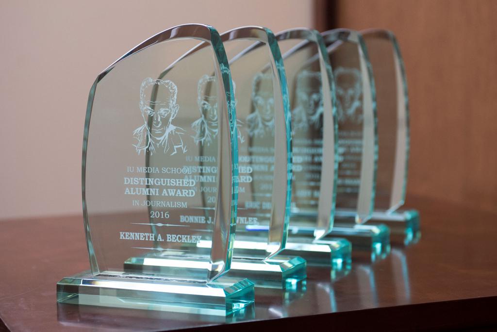 Five Distinguished Alumni Award trophies. The first one is readable. It has an illustration of Ernie Pyle. The text reads: "IU Media School Distinguished Alumni Award in Journalism 2016: Kenneth A. Beckley"