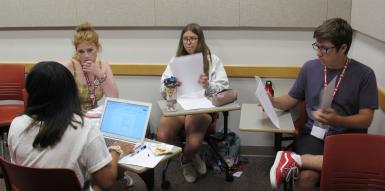 Four students sit at desks in a circle, looking at laptops and papers.