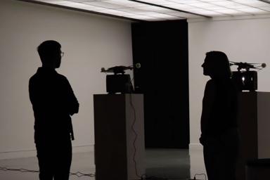 Two people in a darkened room standing in front of two film projectors on pedestals.