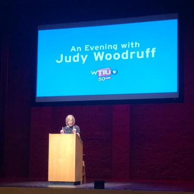 Judy Woodruff speaks at a podium below a sign that says "An Evening with Judy Woodruff."