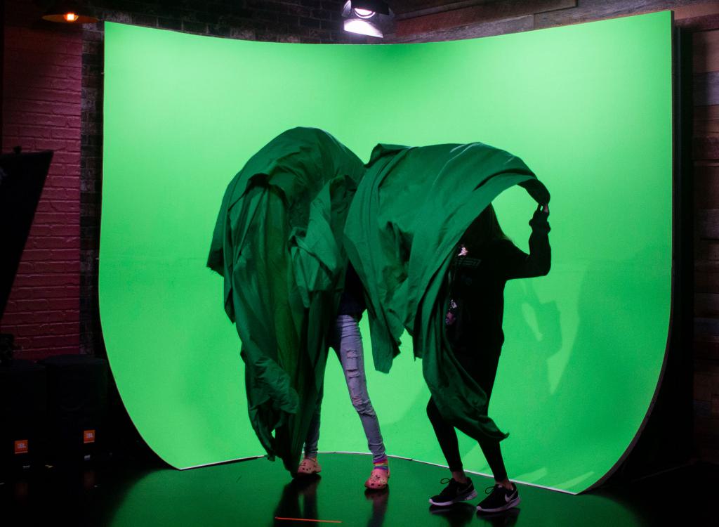 Two girls hide behind green sheets while standing in front of a green screen.