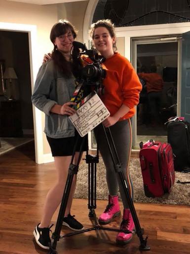 Katie Crump and Riley Dismore pose with a clapboard and a film camera on a tripod.