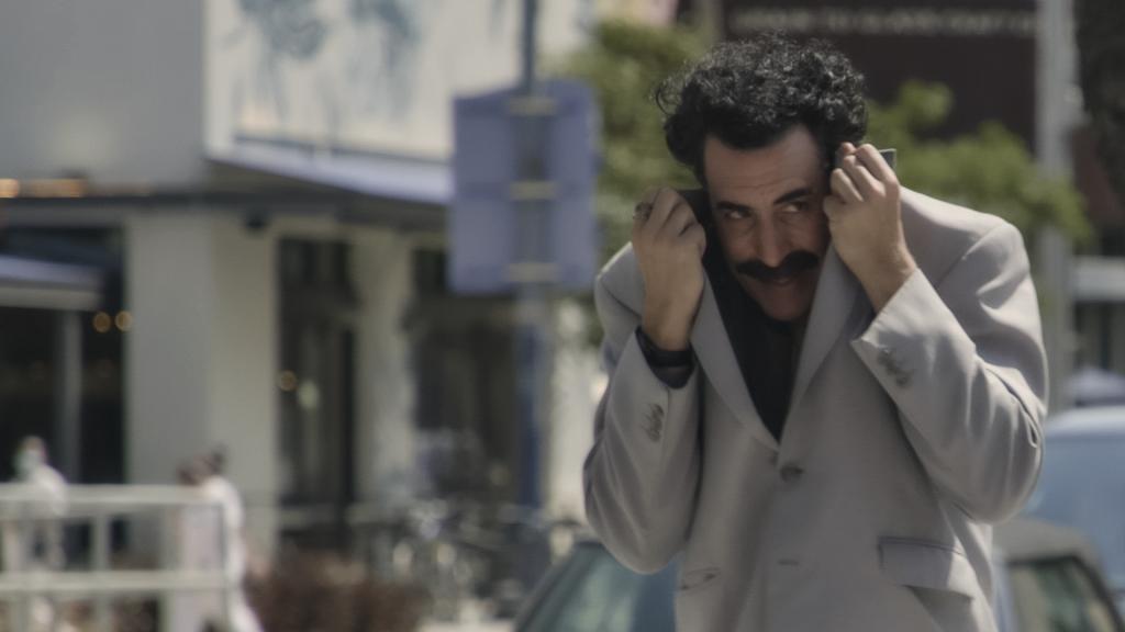 Still image from "Borat Subsequent Moviefilm." Borat is walking down the street covering his face with his coat.