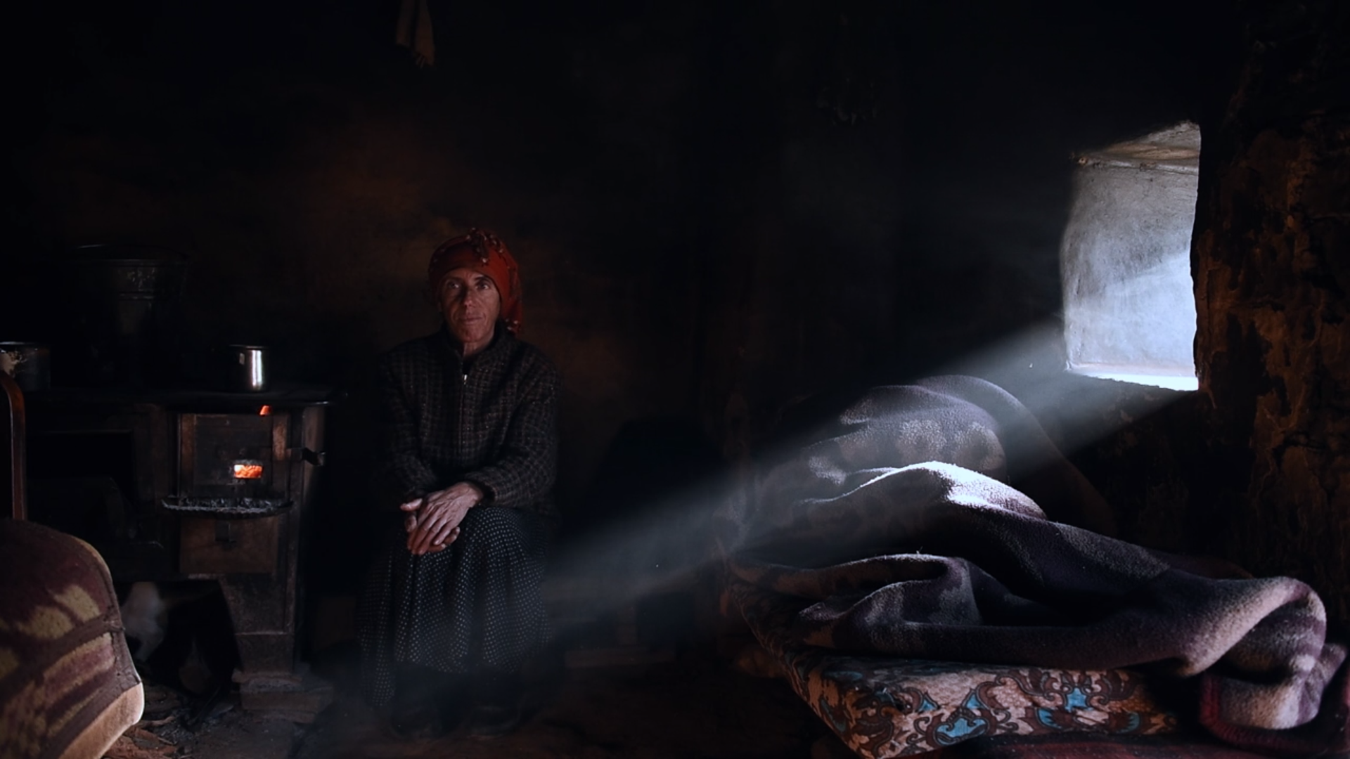 A still image from the movie "Honeyland." A woman sits in a room and watches over her ill mother.