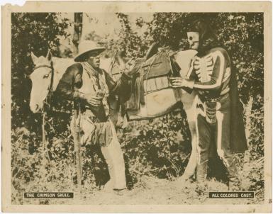 A man wearing a skeleton costume and a man dressed in cowby gear standing in front of a horse.