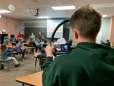 A student in a green sweatshirt films a presentation.