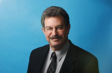 Martin Schram headshot. Man wearing glasses with mustache smiles against blue backdrop.