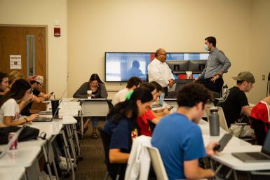 A man talks to a professor in the front of a classroom full of students