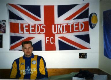 A man in a Leeds United jersey sits in front of a Leeds United flag.