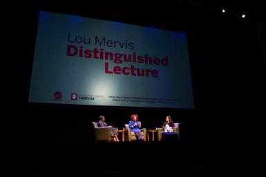 Wide shot of three people on the IU Auditorium stage with a large screen behind them featuring "Lou Mervis Distinguished Lecture" on display 