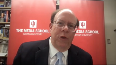 A professor moderates a Zoom conversation while sitting in front of red banners with The Media School logo