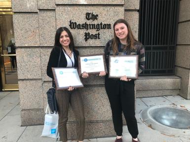 Helen Rummel and Cate Charron stand in front of "The Washington Post" sign displayed on its building. They are holding three framed awards.