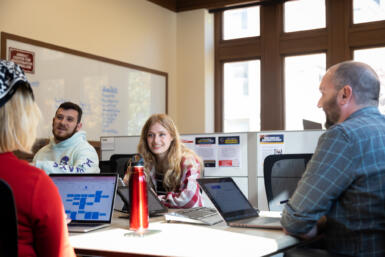 Two students smile as they sit at a table with others working on laptops in the Arnolt Center.