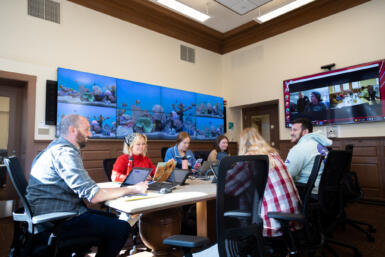 Students and Managing Editor of the Arnolt Center Craig Lyons sit at a table with screens displaying other students on a Zoom call.