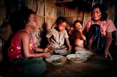 A mother sits next to her three children as they eat food from bowls.