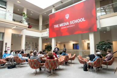 Wide shot of people seated in The Media School commons with Scott Dolson and adjunct professor Les Morris seated in front of a large screen displaying "The Media School: Indiana University"