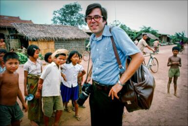 Steve Raymer wears a blue shirt with a bag over his left shoulder with several Cambodian children standing in the background.
