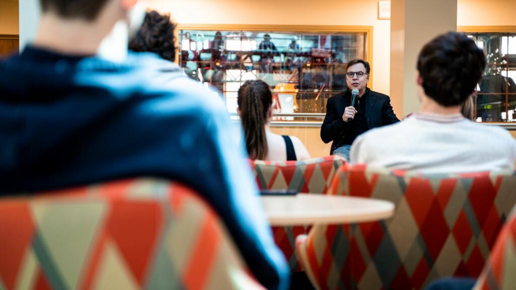Bob Costas speaks into a microphone facing a crowd of students sitting in the Franklin Hall commons.