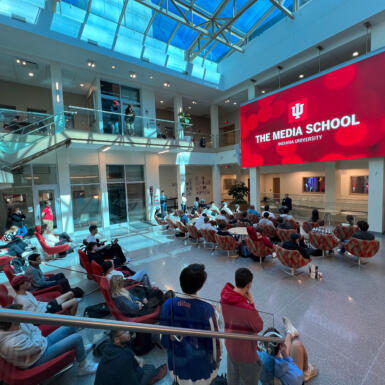 Students sit in chairs listening to Bob Costas sitting in the Franklin Hall commons in front of the large screen that displays "The Media School Indiana University"