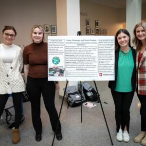 Four students pose for the camera next to their white research poster with black text,