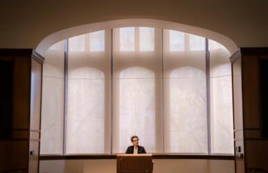 Amy Walter stands in front of large windows in Presidents Hall presenting on stage in front of a podium.
