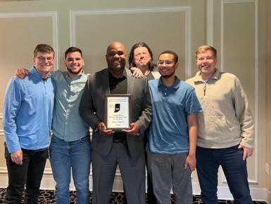 Mike Wells holds an award and stands next to several Media School students and faculty.