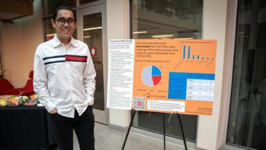 A student smiles into the camera as they stand next to their orange research poster.