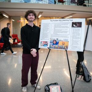 A student with their hands folded in front of them stands next to their white research poster with black text.