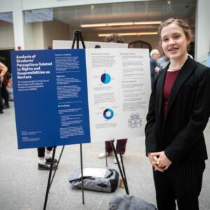 A student in a black blazer, black dress pants, and maroon shirt stands next to their blue-and-white research poster.