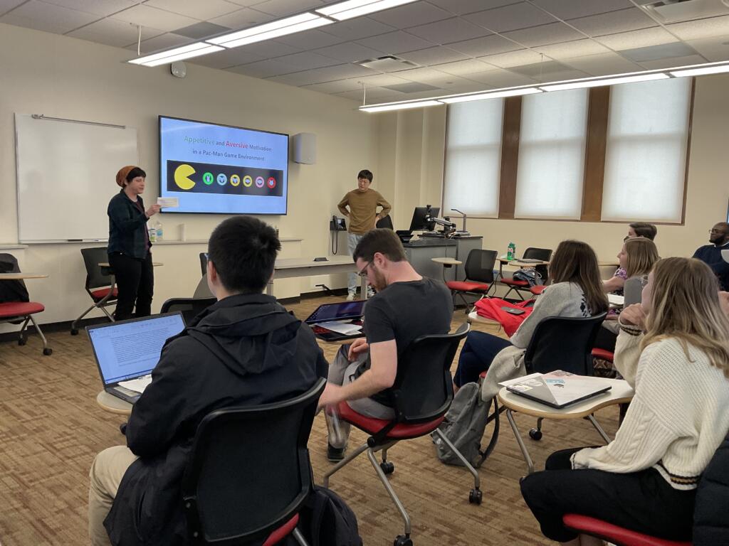 Students sit in a classroom and look at a presentation on a large screen at the front of the room.