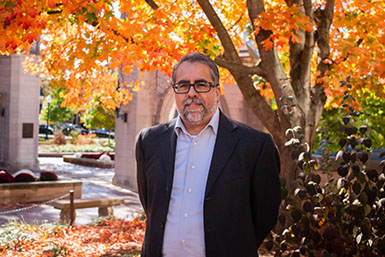 A headshot of Gerry Lanosga standing in front of trees with his arms behind his back