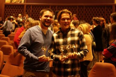 Two people holding several awards stand in a crowd in among the IU Cinema seating.