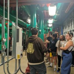 Several students stand around green pipes in a basement.