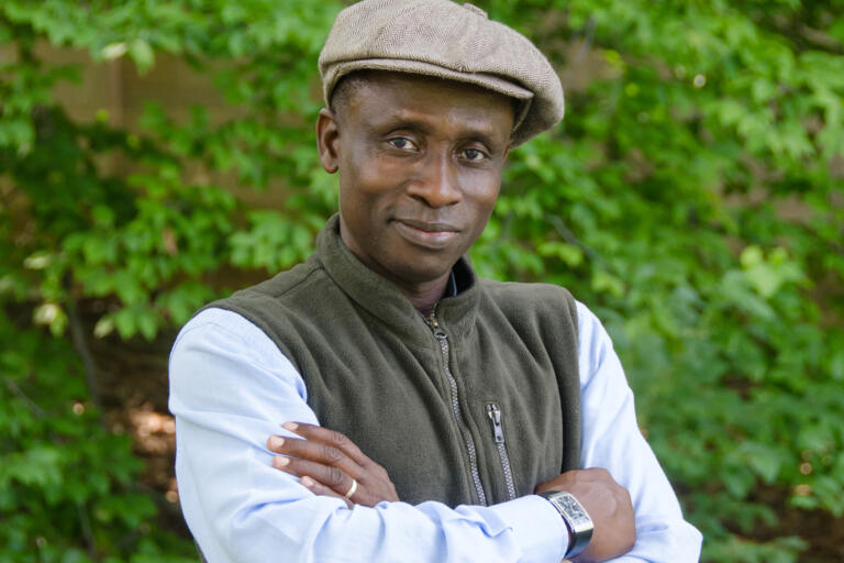 Akin Adesokan stands for a headshot with his arms crossed in front of greenery.
