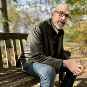 Photo of David Tolchinsky sitting on a bench outside with greenery behind him.