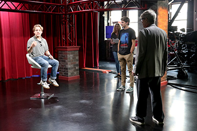 A student sits in a chair in the middle of the room, watched by Eric Roberts and Tristan Reed.