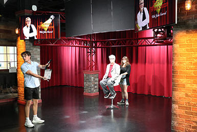 A student adjusts Eric Roberts' chair in the middle of the room while another student holds a clapperboard ready to start a take.