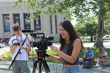 A student adjusts a camera on a tripod, with another student holding headphones in the background.